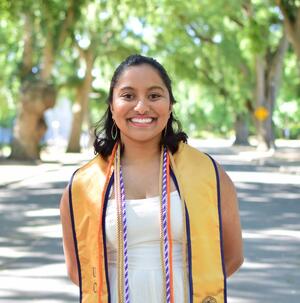 Indira smiling with a graduation sash and cords around her neck
