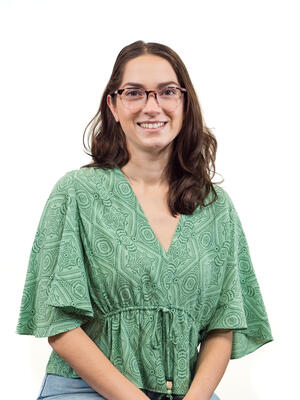 A smiling white woman with brown hair and glasses, wearing a teal green patterned shirt and blue jeans, stands in front of a white background.